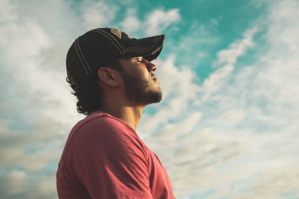 Man in red shirt and black cap, looking peaceful with eyes closed and lovely sky behind him. 